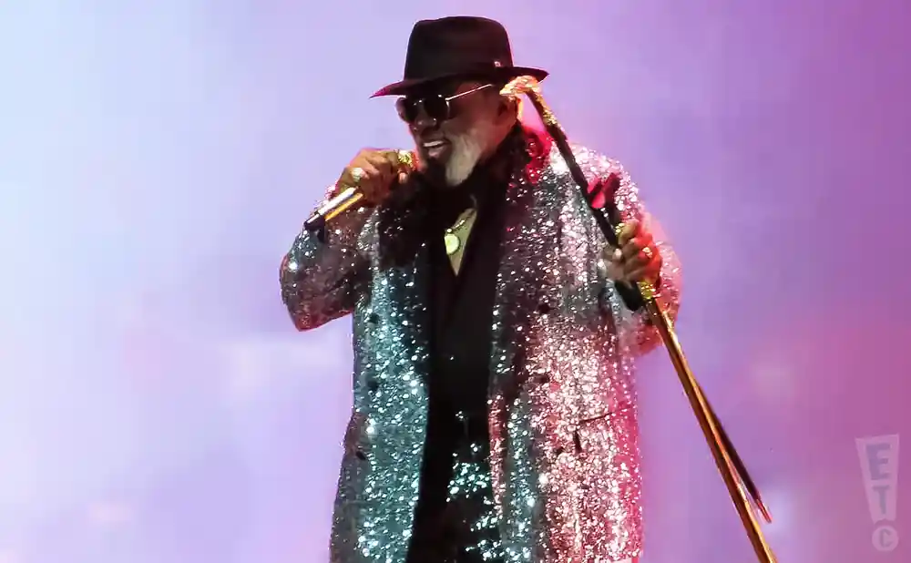 A man in a sparkly jacket and fedora sings into a microphone while holding a cane.