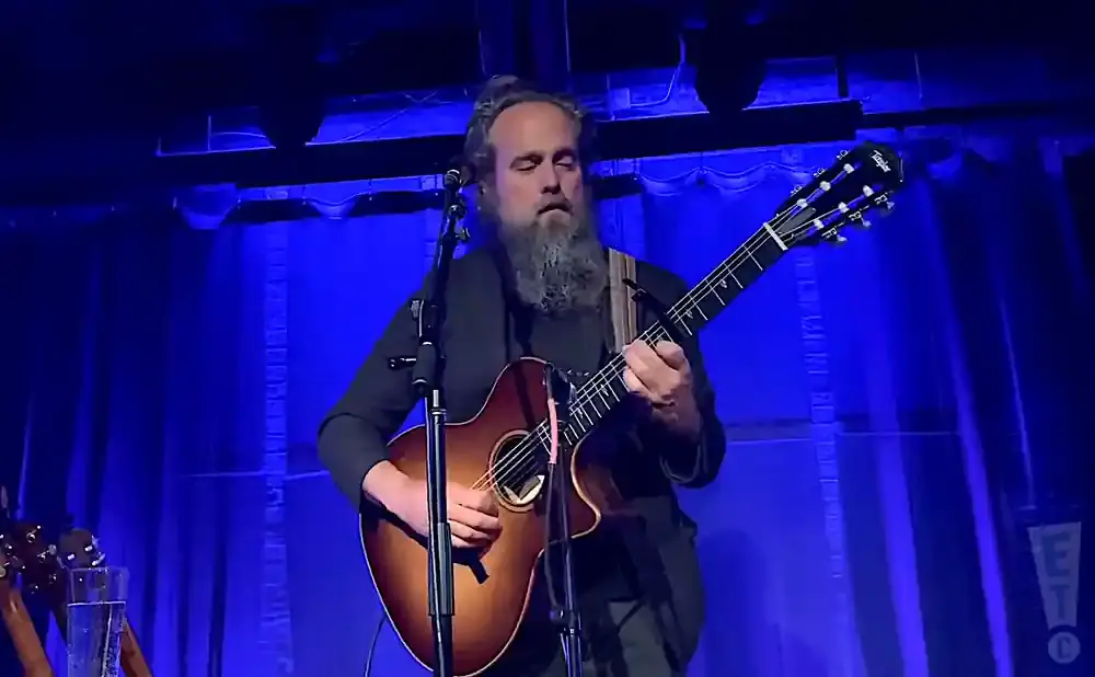 A man with a long beard plays an acoustic guitar on a blue-lit stage.