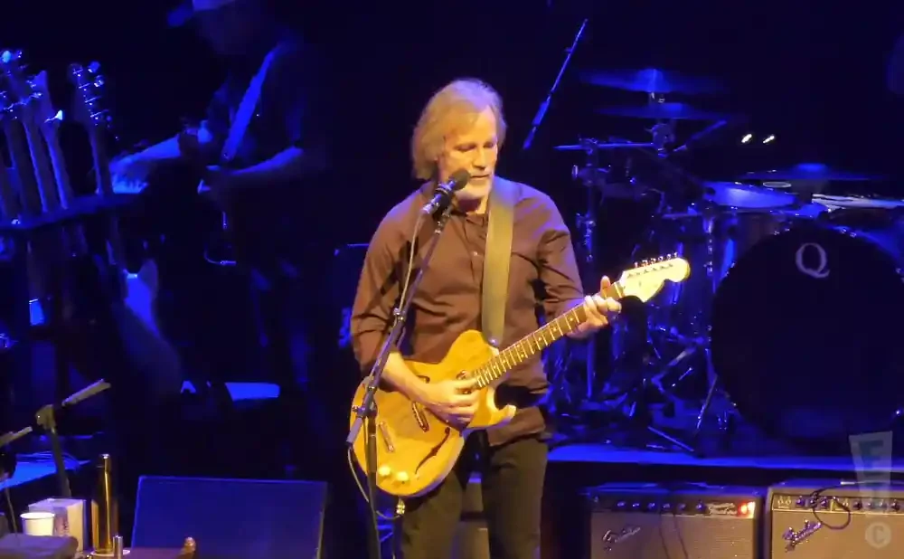 A man with long, graying hair plays a guitar on stage. He wears a brown shirt and black pants.