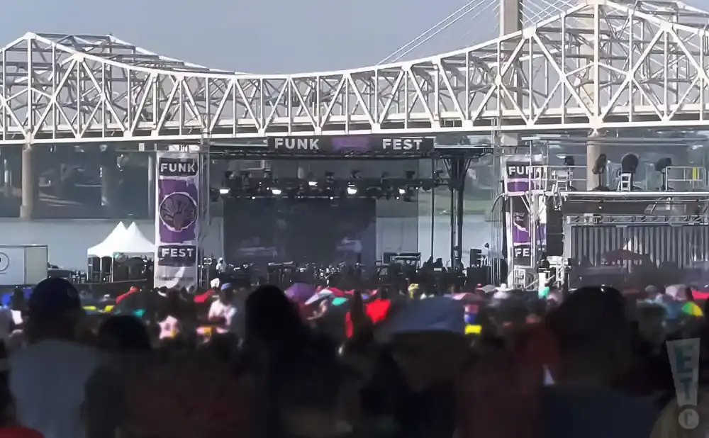 A crowd watches a stage at an outdoor festival under a large metal bridge. Banners read 
