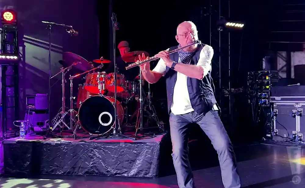 Man playing a flute on stage with drums and lighting.