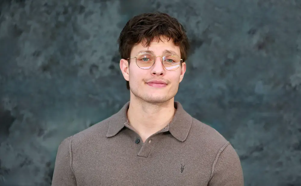 A man with glasses and a brown polo shirt smiles against a dark, textured background.