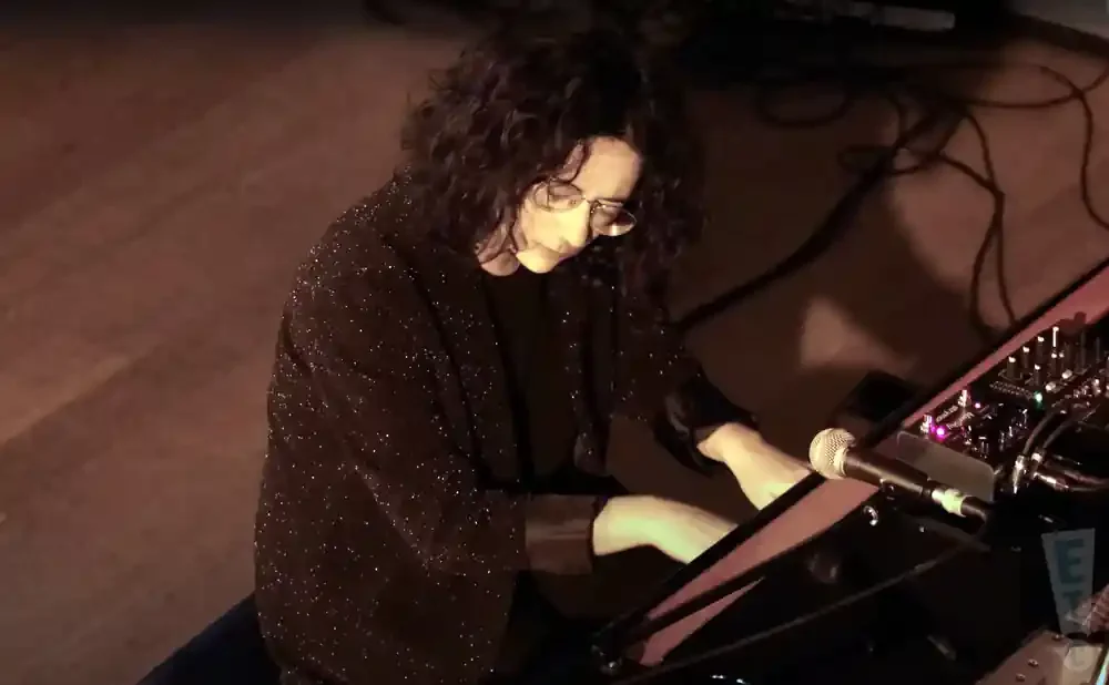 A woman with curly dark hair and glasses plays a keyboard, wearing a sparkly black jacket.