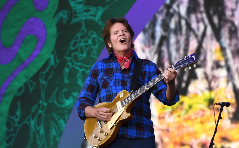 Guitarist John Fogerty performs on stage wearing a blue plaid shirt and red bandana.