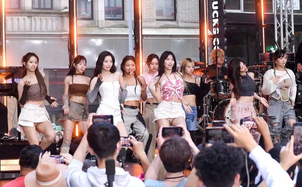 A group of young women perform on stage, with fans holding up phones to record.