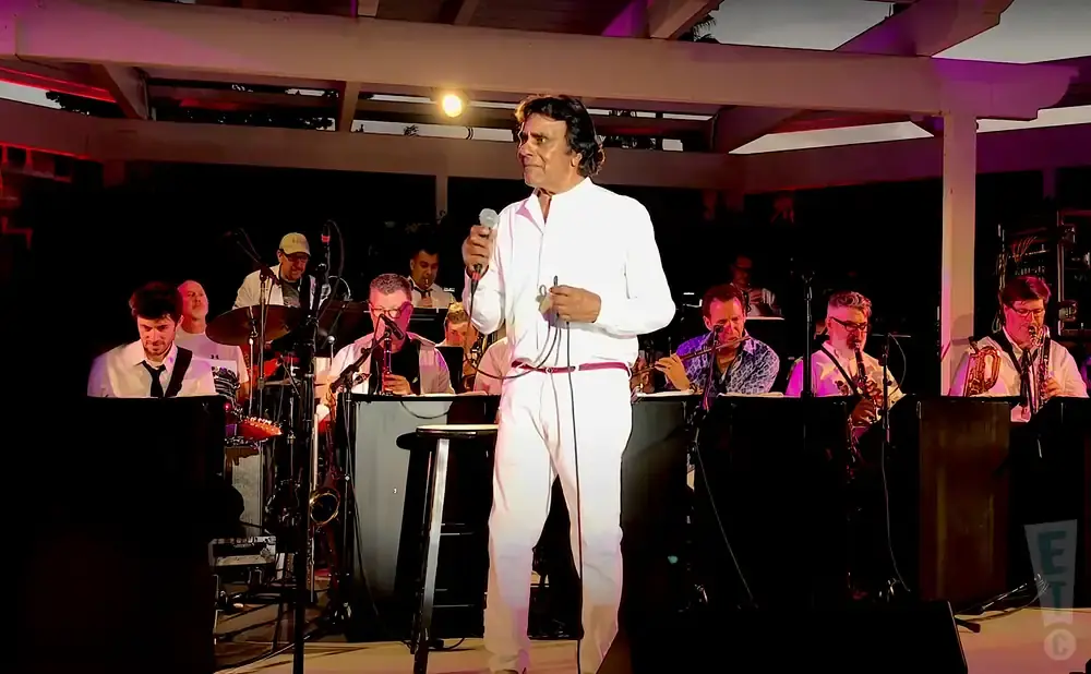 A male singer in a white suit performs with a band playing various instruments.