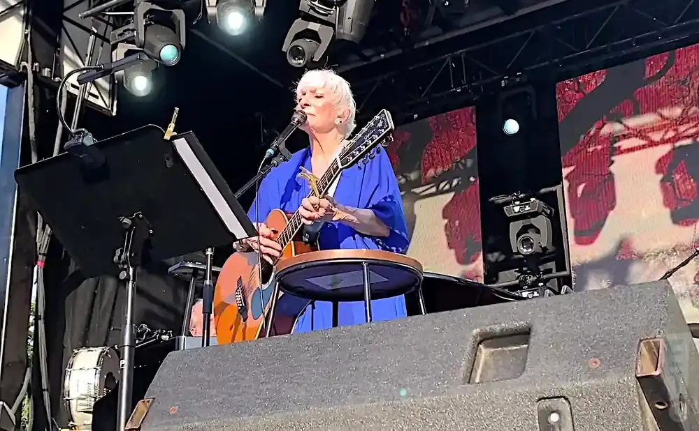 Singer with white hair plays an acoustic guitar on stage, wearing a blue outfit.