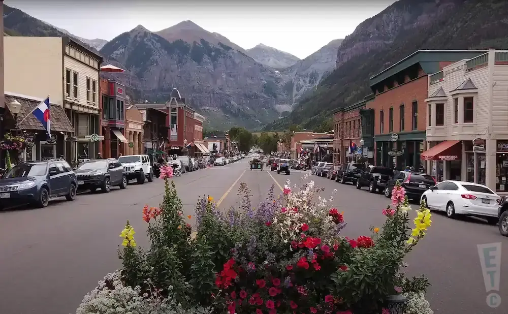 Street view of a town in a mountain valley, with buildings on either side and cars parked along the road. Flowers are in the foreground.