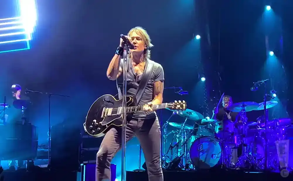 A man plays an electric guitar and sings into a microphone on a stage with blue lighting.