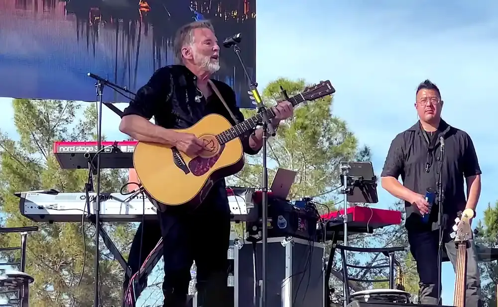 A man with a beard plays an acoustic guitar on stage, with another man standing behind him holding a bass guitar.