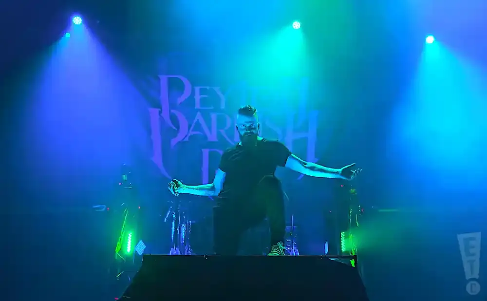 A man with a beard and a black t-shirt performs on stage with blue and green lights behind him.