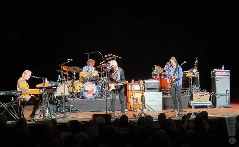 A band performs on stage, with a keyboardist on the left, drummers in the middle, and guitarists on the right.