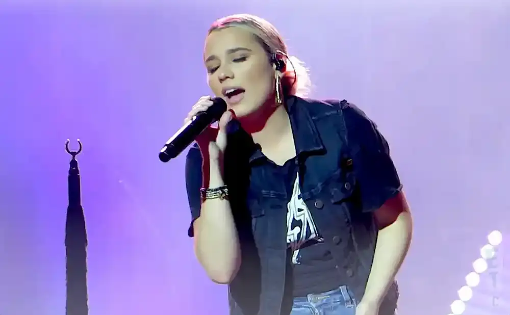 Singer Gabby Barrett performs on stage, holding a microphone and wearing a denim vest over a t-shirt.
