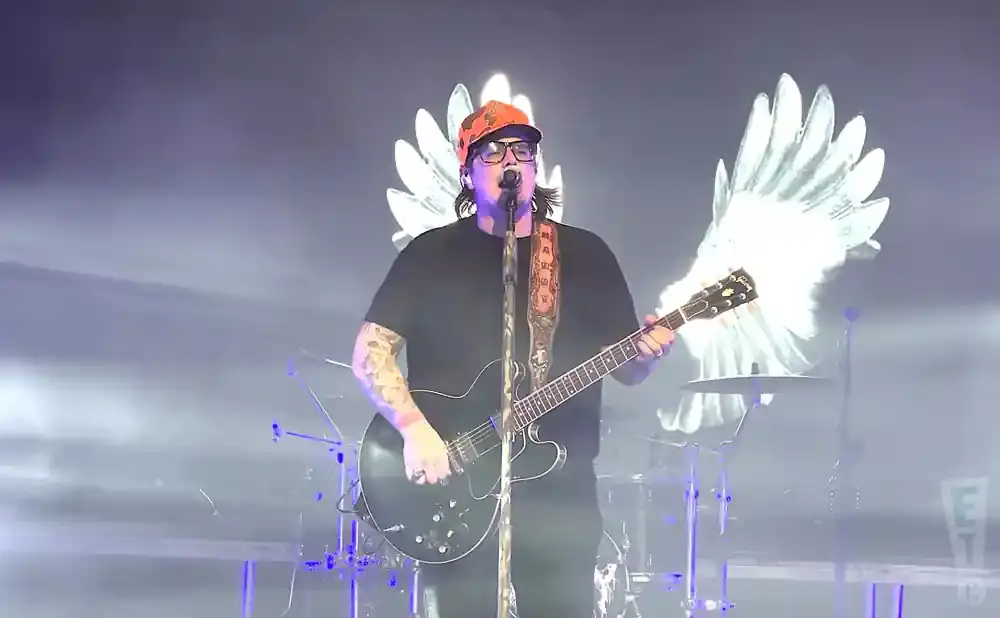 A man in a black shirt and orange hat plays an electric guitar on stage with glowing white wings behind him.