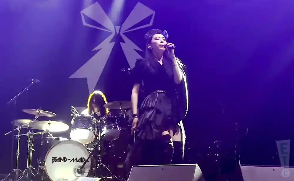 A female singer performs on stage with a drummer and a large white ribbon logo in the background.