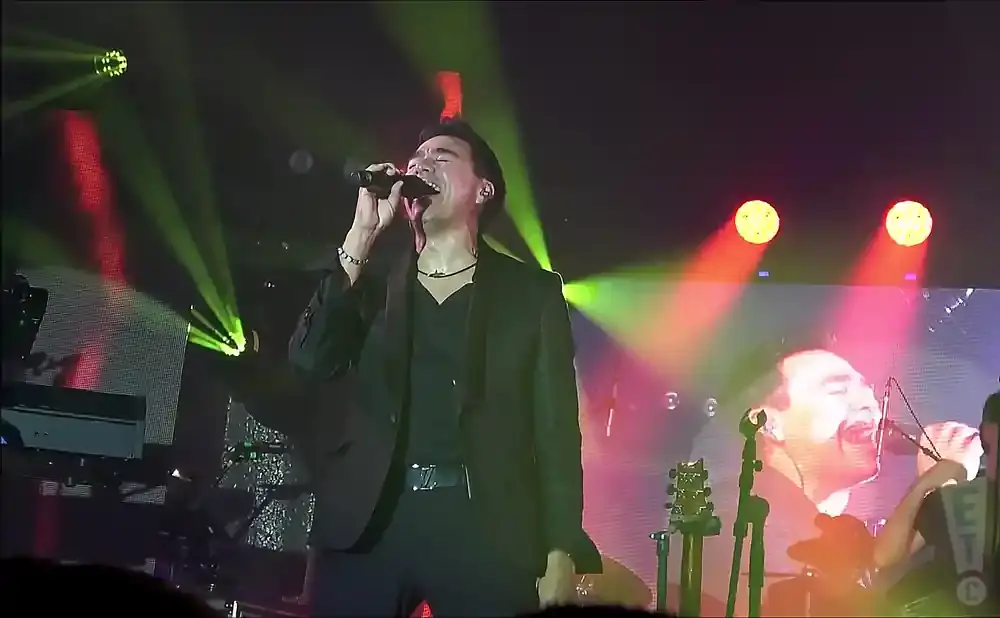 A male singer in a black suit and V-neck shirt sings into a microphone on stage with bright lights.