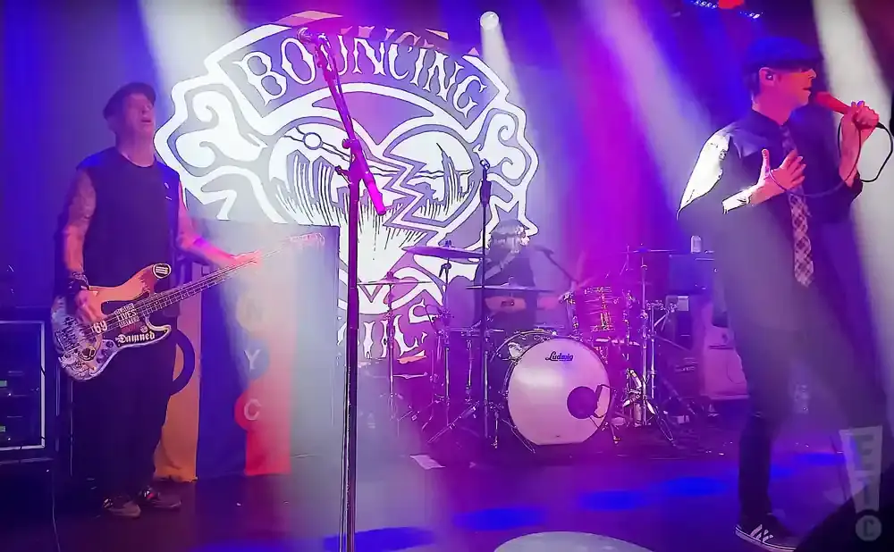 A band performs on stage with purple and white lighting. The bassist is on the left, drummer in the center, and singer on the right.