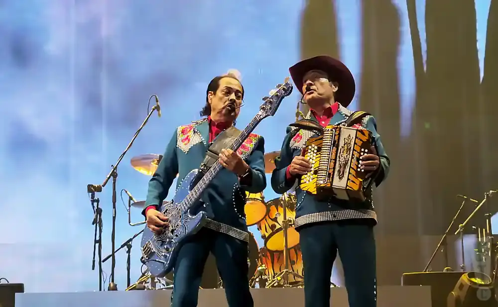 Two men on stage in matching teal suits with ornate collars play music: one plays a blue bass guitar, the other an accordion.