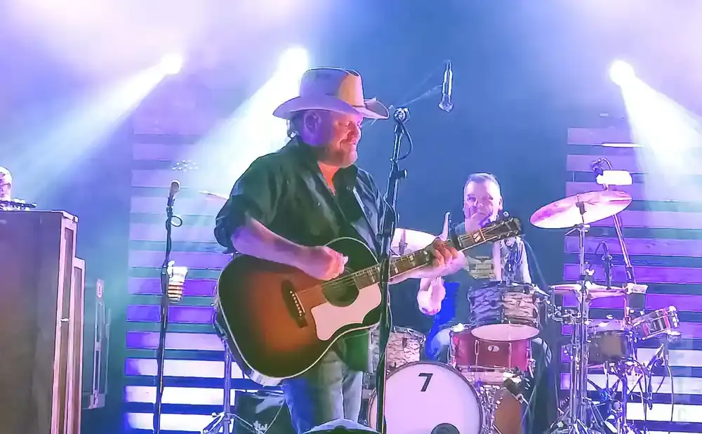 A man in a cowboy hat plays an acoustic guitar on stage with a drummer behind him.