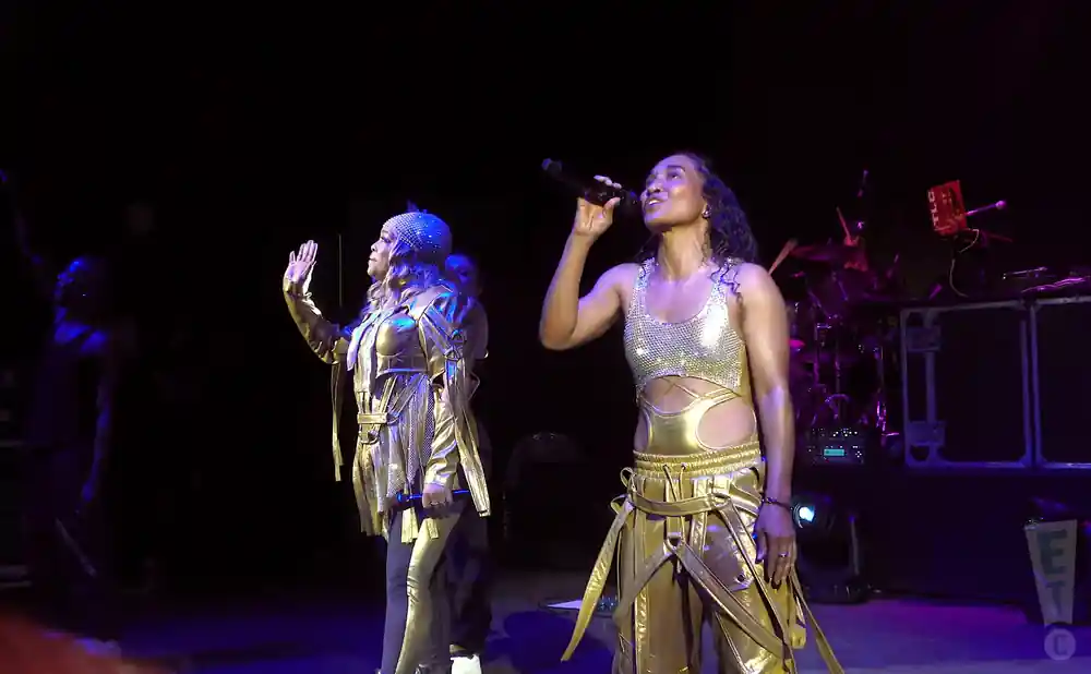 Two women in gold outfits perform on stage, one singing into a microphone, the other waving.