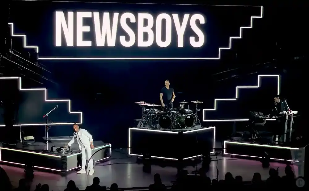 Newsboys band performs on stage with a large illuminated sign and tiered, neon-lit platforms.