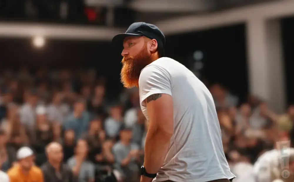 Man with a red beard wearing a black cap and white t-shirt, seen from the side, with a blurred audience behind him.