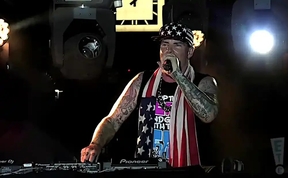 A man wearing an American flag hat and scarf raps into a microphone while standing behind a DJ booth.