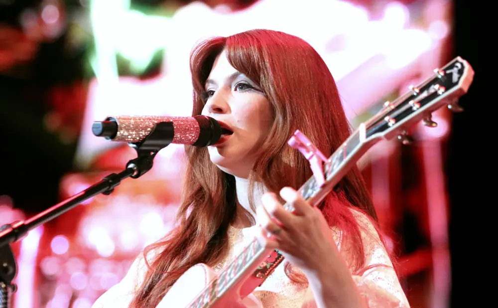 A woman with red hair sings into a sparkly microphone while playing a pink guitar on stage with colorful lights.