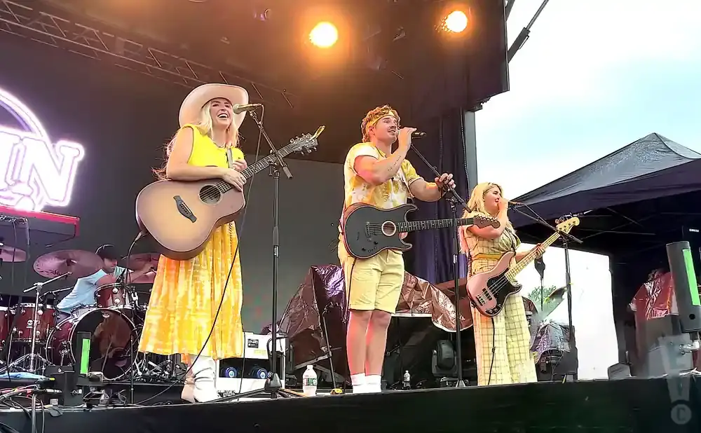A band performs on stage under bright lights, with singers playing guitar and bass.