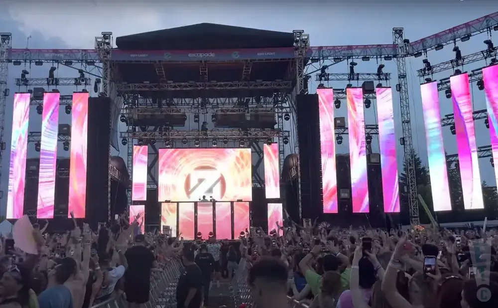 An outdoor music festival stage with bright LED screens and a large crowd.