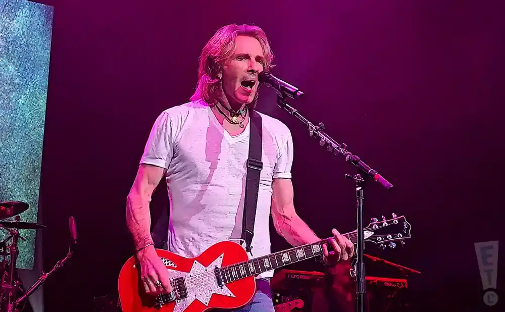 Musician playing a red electric guitar with a white star on a stage with pink lighting.