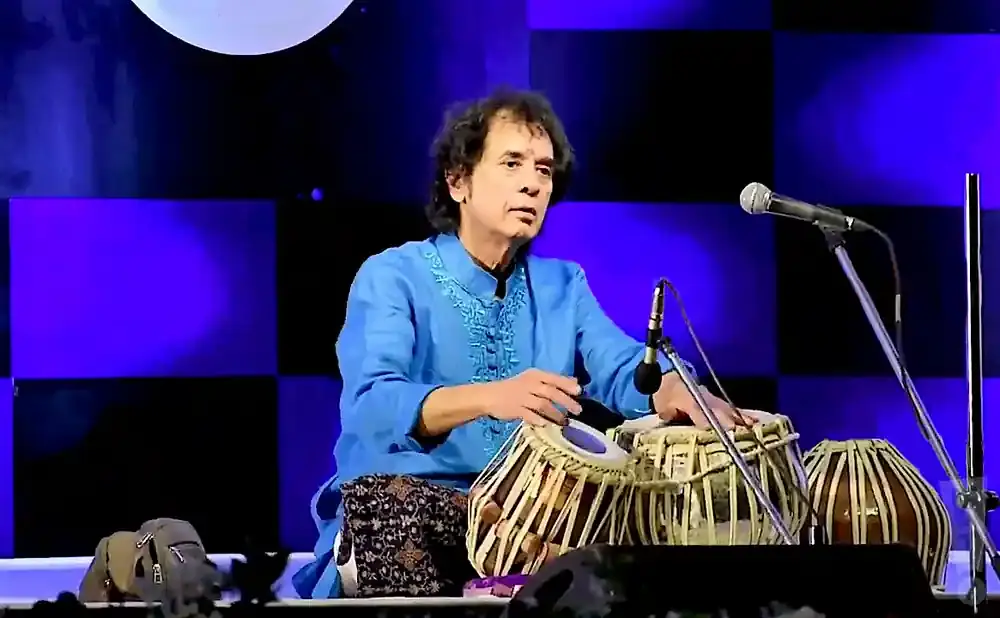 A man in a blue tunic plays the tabla drums in front of a blue checkered backdrop.