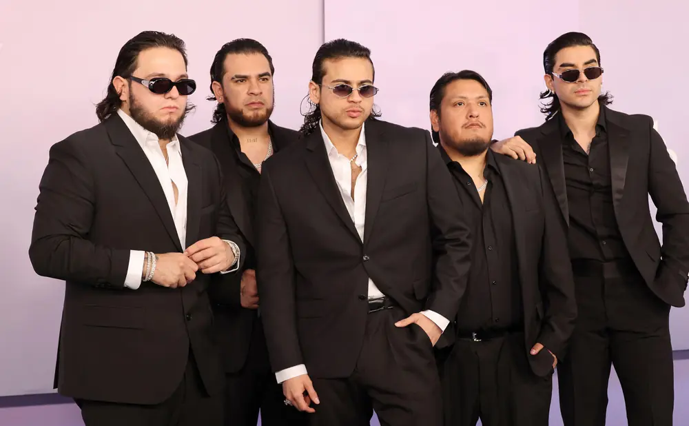 Five men in black suits and sunglasses pose for a photo against a light purple background.