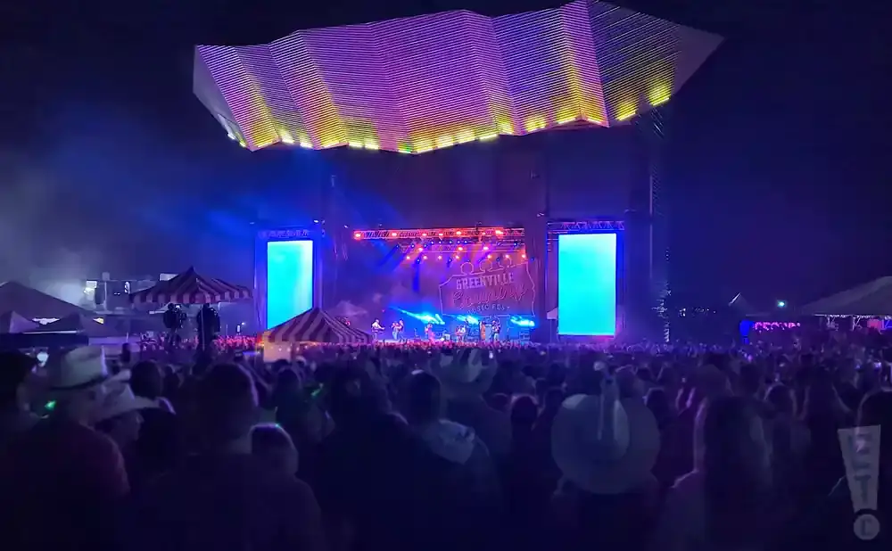 A large crowd watches a band perform on a brightly lit stage at the Greenville Country Music Fest at night.