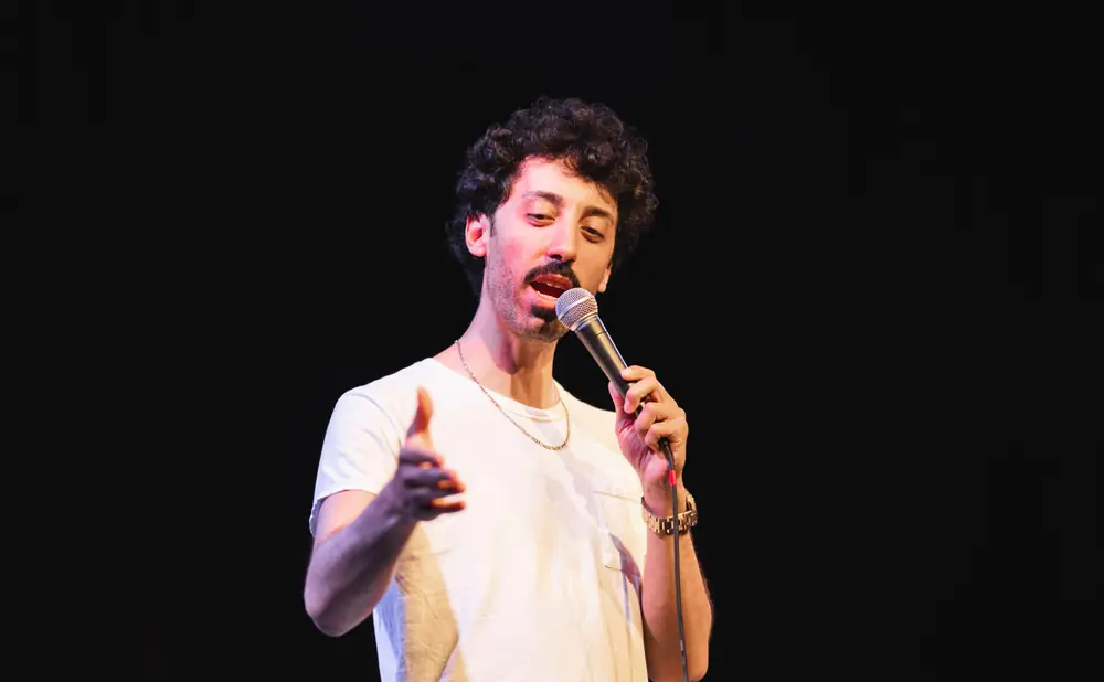 A man with curly hair and a mustache speaks into a microphone on a dark stage.