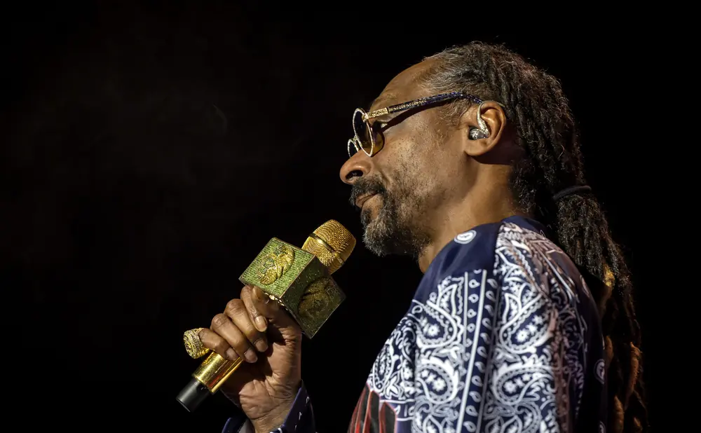 Snoop Dogg on stage holding a gold microphone with diamond accents, wearing glasses and a bandana print shirt.