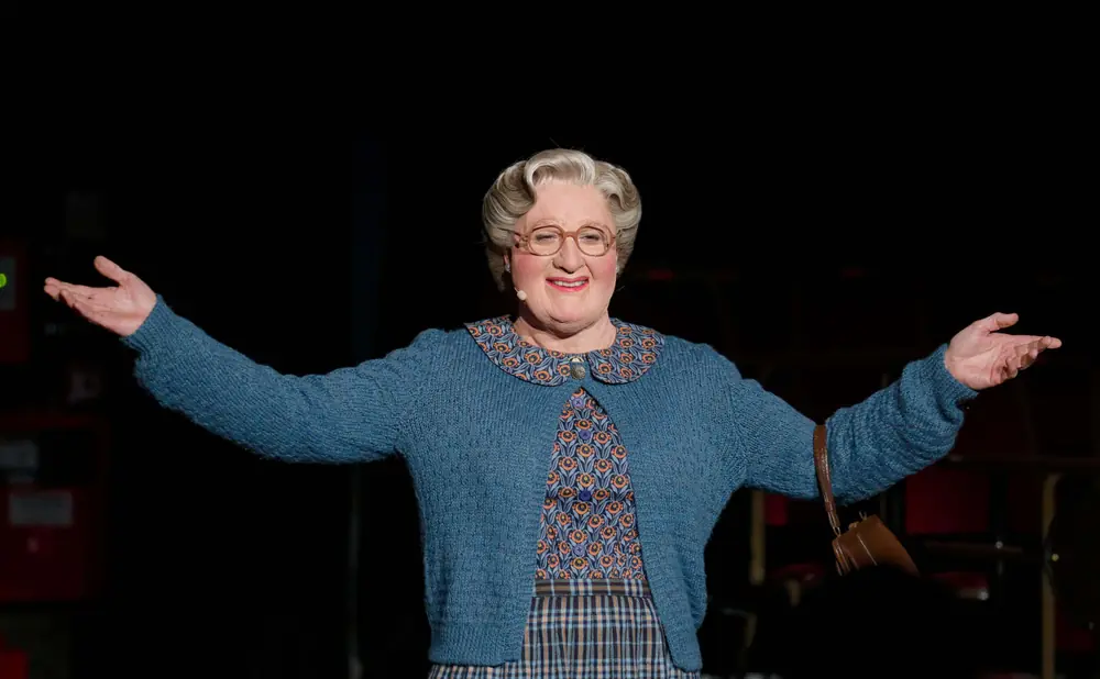 Woman dressed as Mrs. Doubtfire with arms outstretched, wearing a blue cardigan and patterned dress.