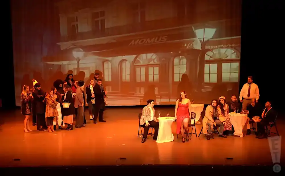A group of actors in period costume are on a stage in front of a projected backdrop of a building.