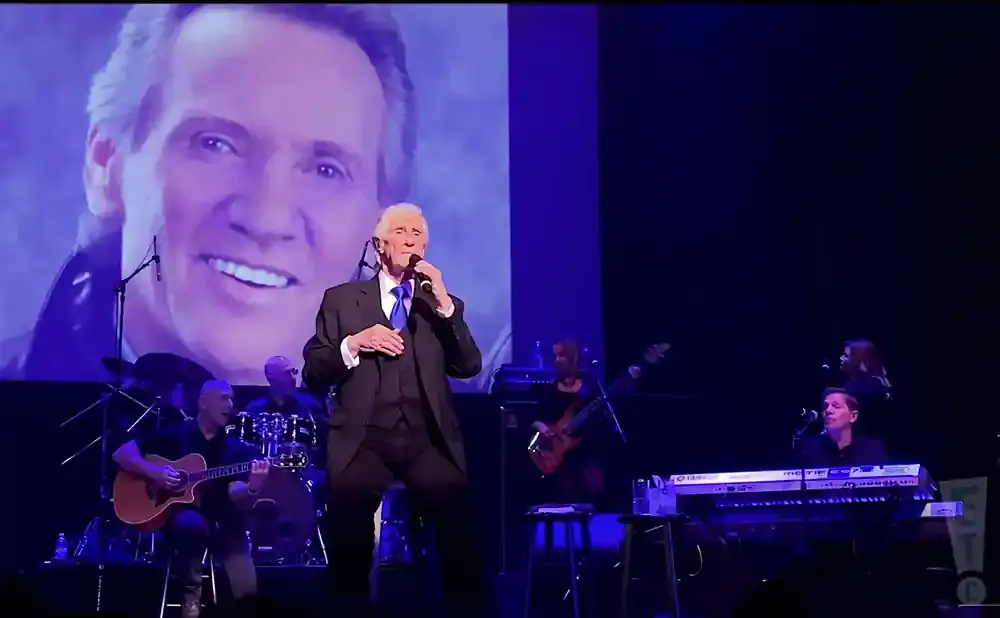 A performer in a suit sings on stage with a projection of his face behind him.