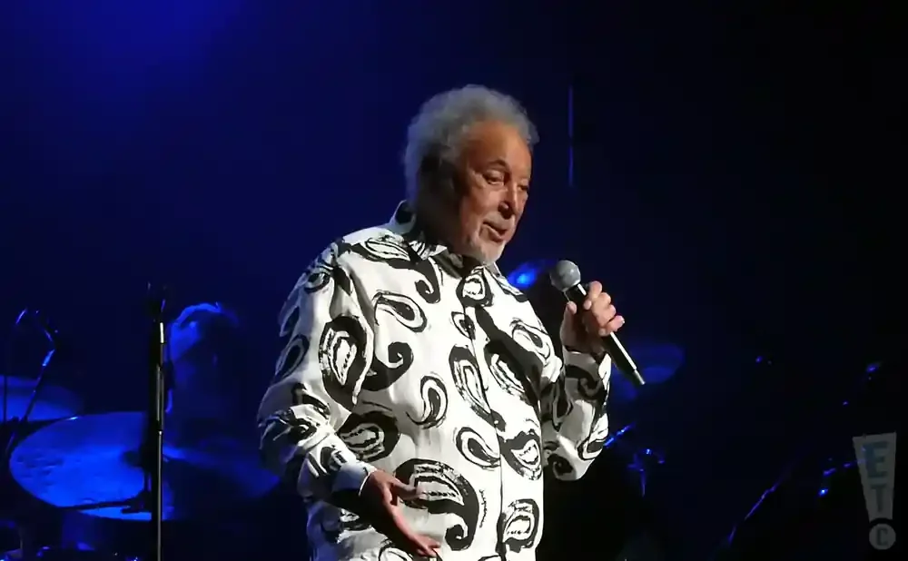 Sir Tom Jones performs on stage wearing a black and white patterned shirt, holding a microphone.