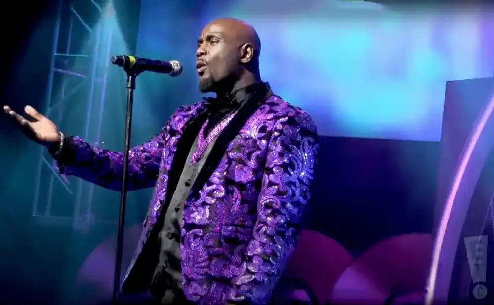 A bald Black man in a sequined purple jacket sings into a microphone on a stage.