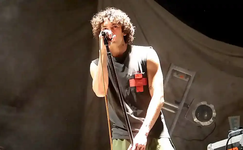 A young man with curly hair sings into a microphone on stage, wearing a sleeveless shirt with a red cross emblem.