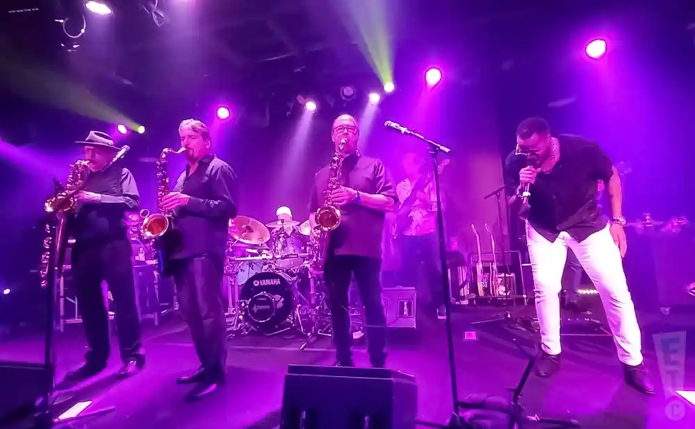 A band plays on a stage with purple lighting. Four men are visible: three playing saxophones and one singing into a microphone.