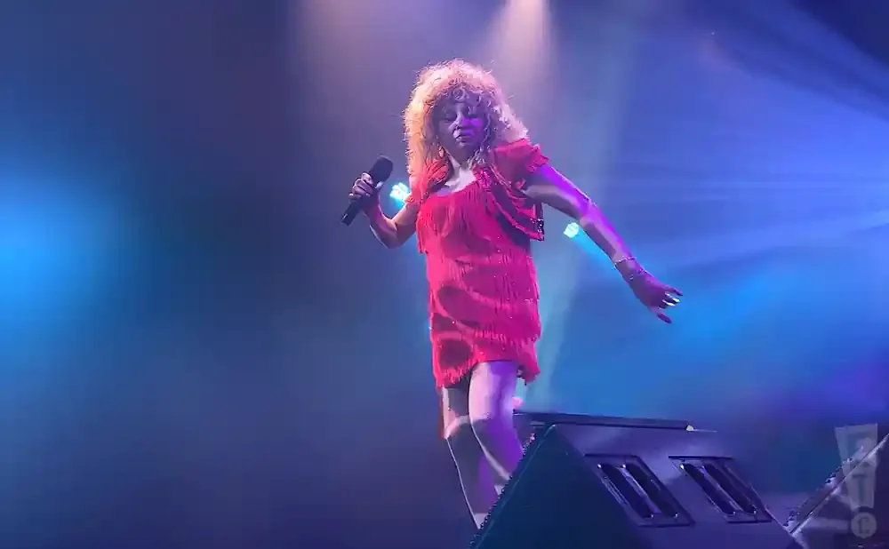 A woman in a red fringed dress and jacket sings into a microphone on a stage with blue lighting.
