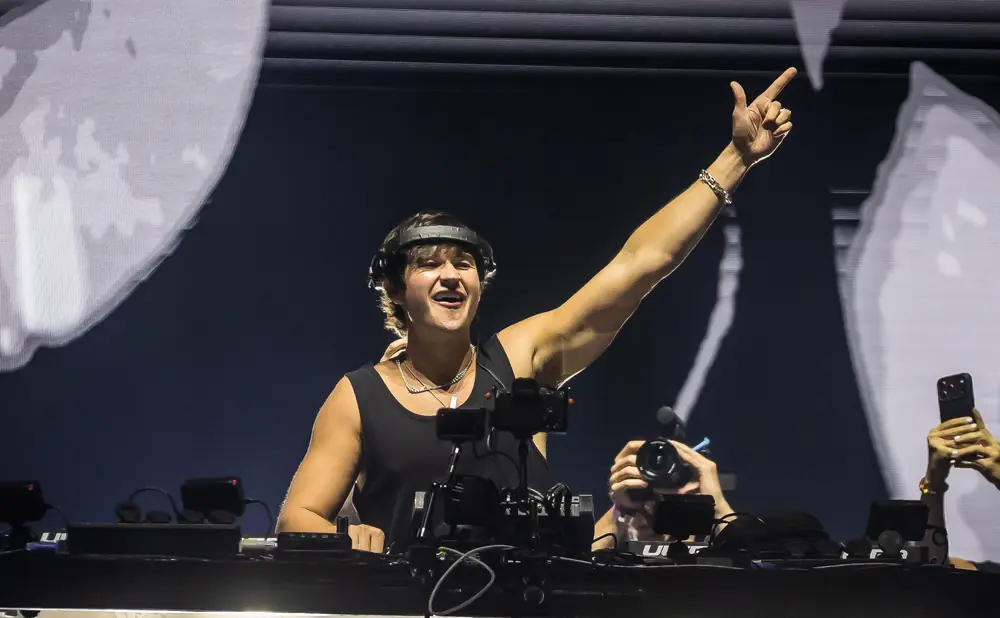 DJ raising a hand in a club with headphones and a necklace.