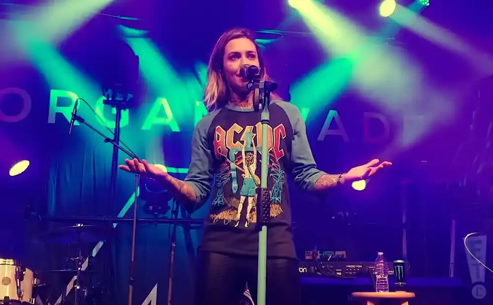A female singer performs on stage wearing an AC/DC shirt, with colorful lights and a band name on the backdrop.