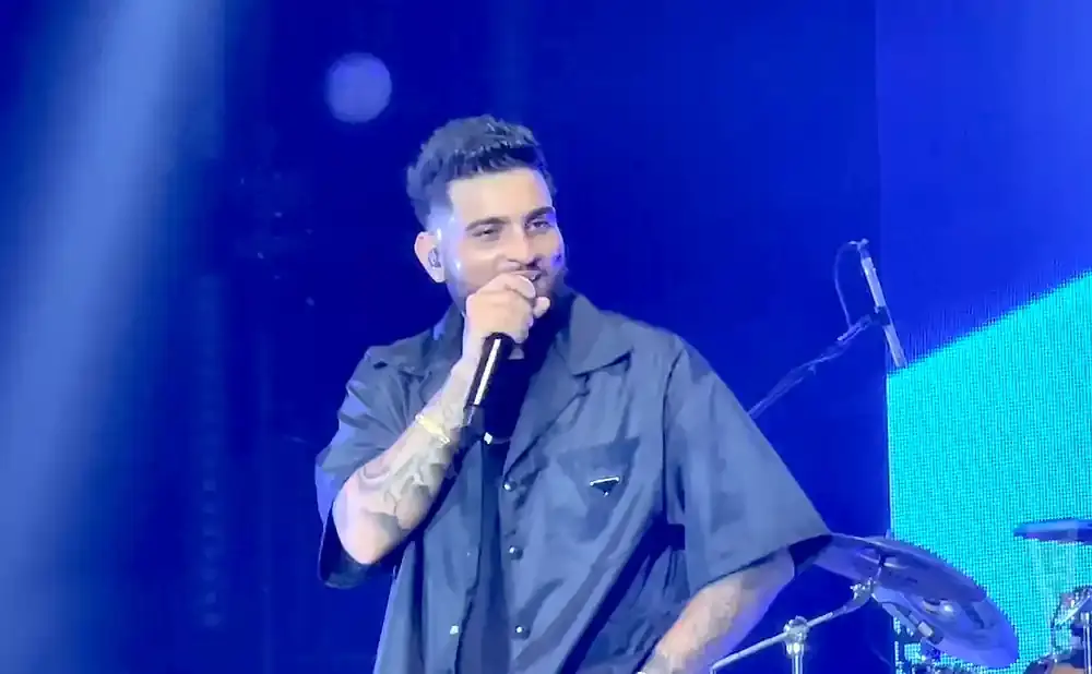 A man in a dark open-collar shirt performs on stage with a microphone, illuminated by blue lights.