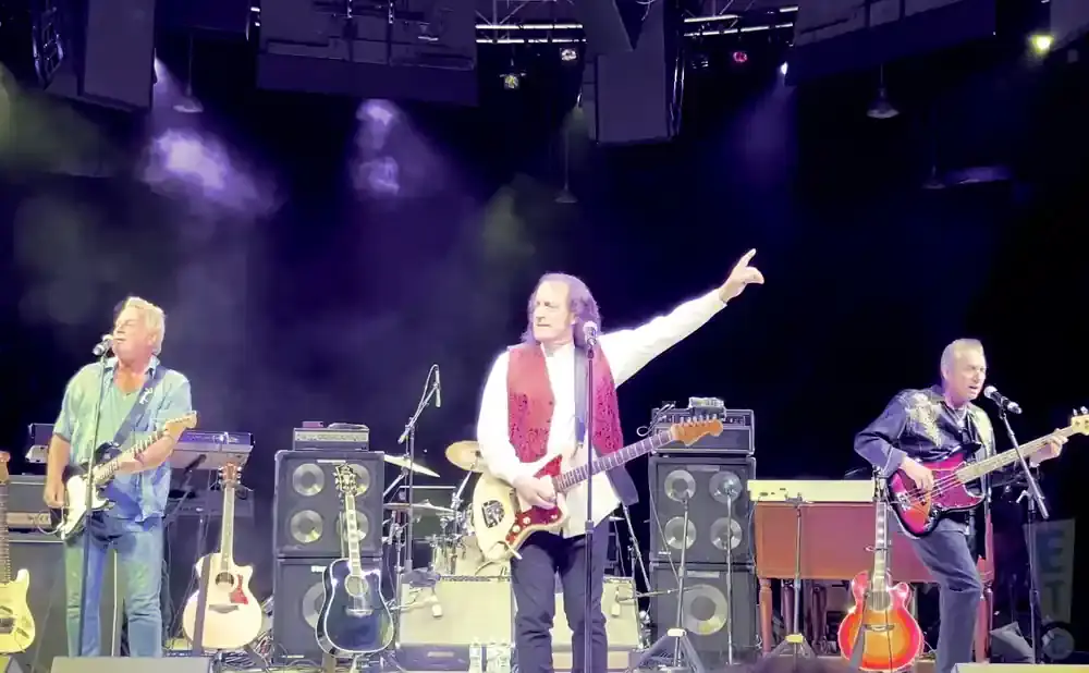 Three musicians perform on stage: one plays guitar and sings, another plays guitar, and a third plays bass guitar.