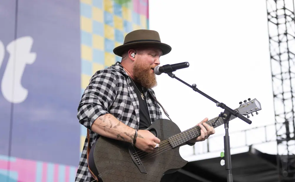 A bearded man in a fedora and checkered shirt plays an acoustic guitar on stage, singing into a microphone.