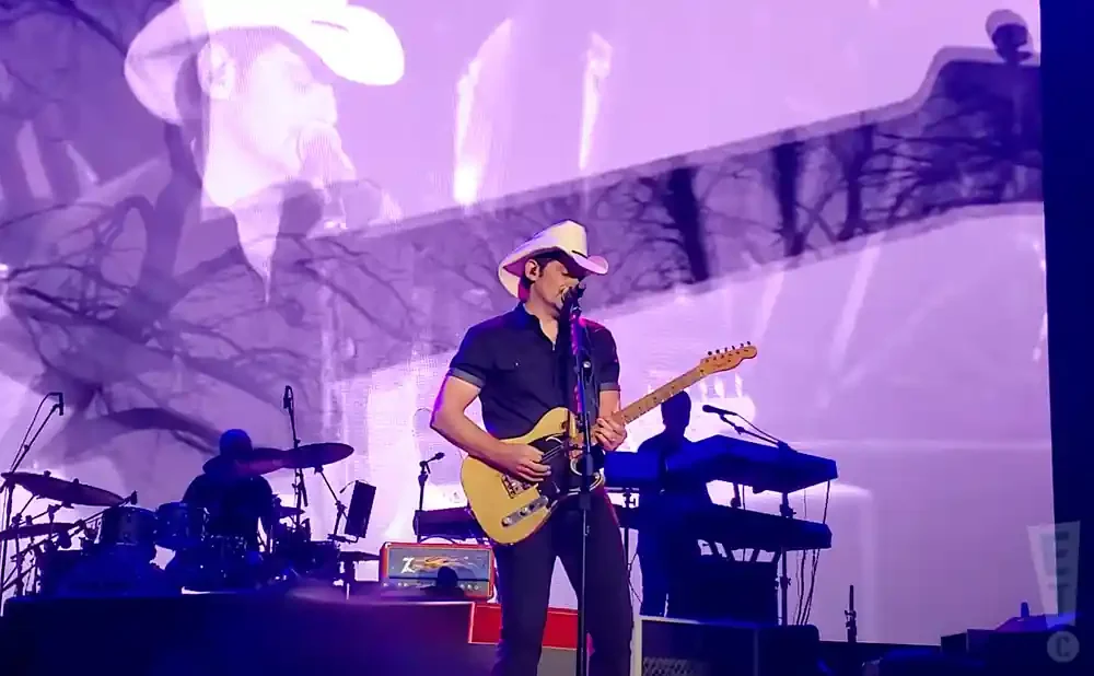 A male singer in a cowboy hat plays guitar on stage with a drummer and keyboardist behind him.